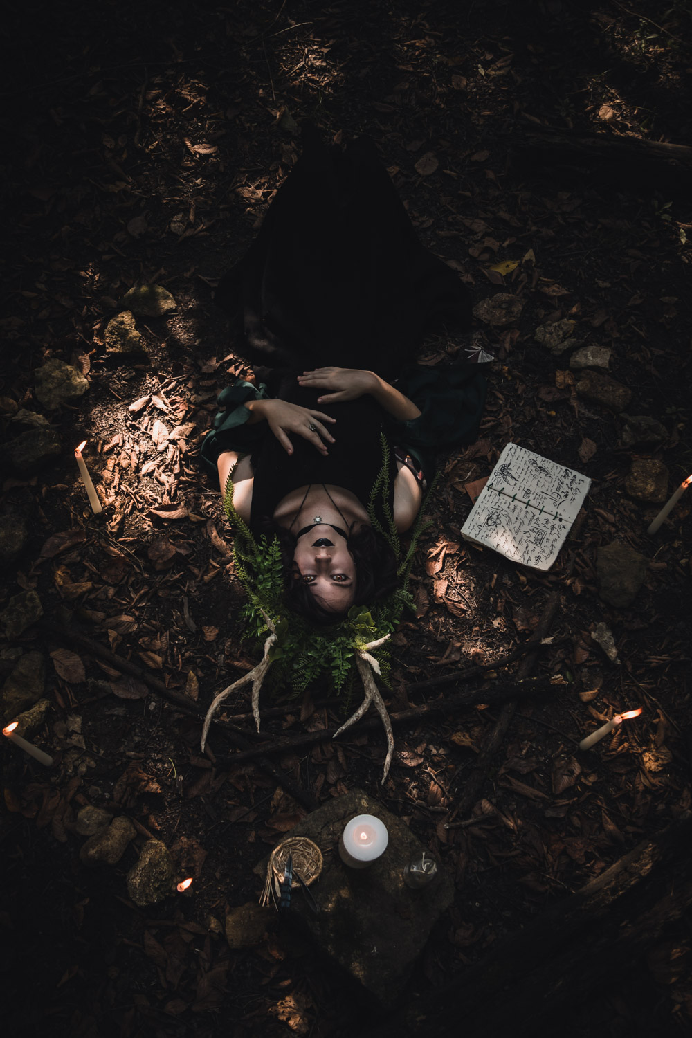 Fine Art Photograph of a woman in a black witch dress and ornate vine and leaf headpiece lying on the leaf covered ground surrounded by spellbooks and candles